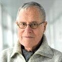 Ein Mann mit Brille trägt eine Strickjacke und einen Schal. Er lächelt in die Kamera.