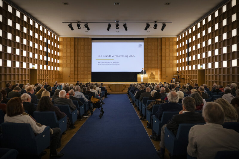 Männer und Frauen sitzen in einem großen Saal. Auf einem Bildschirm ist der Titel der Veranstaltung eingeblendet: Leo-Brandt-Veranstaltung 2025