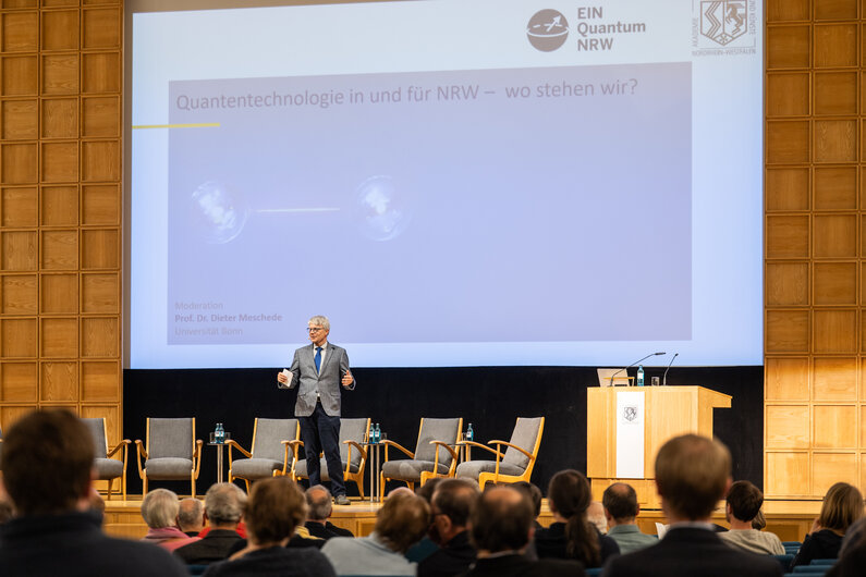 Ein Mann steht auf einer Bühne. Im Hintergrund ist der Titel der Veranstaltung "Quantentechnologie in und für NRW – wo stehen wir?" eingeblendet. 