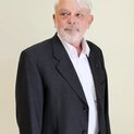 Ein Mann mit Vollbart steht vor einer Wand und schaut nach links. Er trägt eine Anzugjacke.