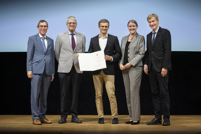 Vier Männer und eine Frau haben sich auf einer Bühne für ein Foto aufgestellt. Der junge Mann in der Mitte hält eine Urkunde in den Händen.