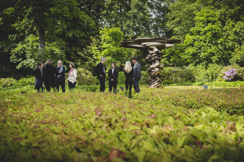 In einem Garten stehen Männer und Frauen in kleinen Gruppen zusammen und unterhalten sich.