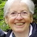Eine Frau mit Brille steht vor Sträuchern und lächelt strahlend in die Kamera.