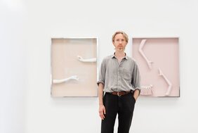 Der Bildhauer Andreas Schmitten steht vor einer weißen Wand. Links und rechts von ihm hängen zwei seiner Werke.  