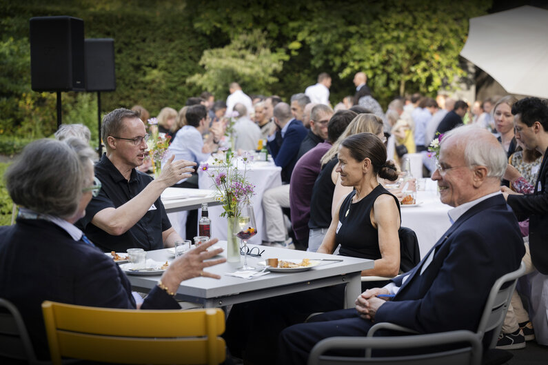 Männer und Frauen sitzen im Garten der Akademie an Tischen.