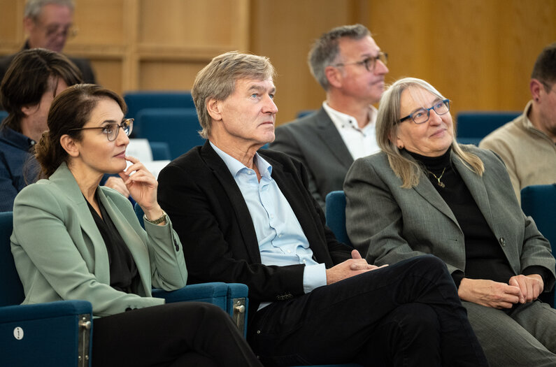 Männer und Frauen sitzen in einem Veranstaltungsraum auf blauen Stühlen.
