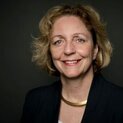 Eine Frau lächelt in die Kamera. Sie trägt einen Blazer und eine goldene Halskette.