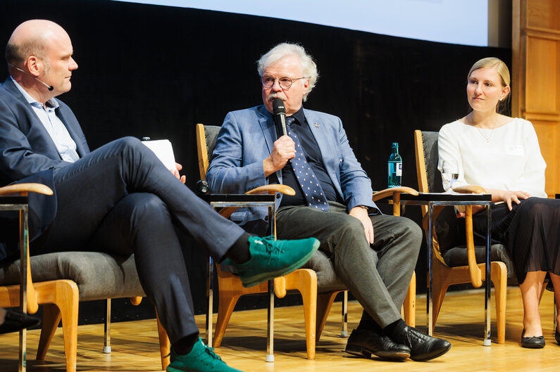 Zwei Männer und eine Frau sitzen auf einem Podium. Einer der Männer spricht in ein Mikrofon.