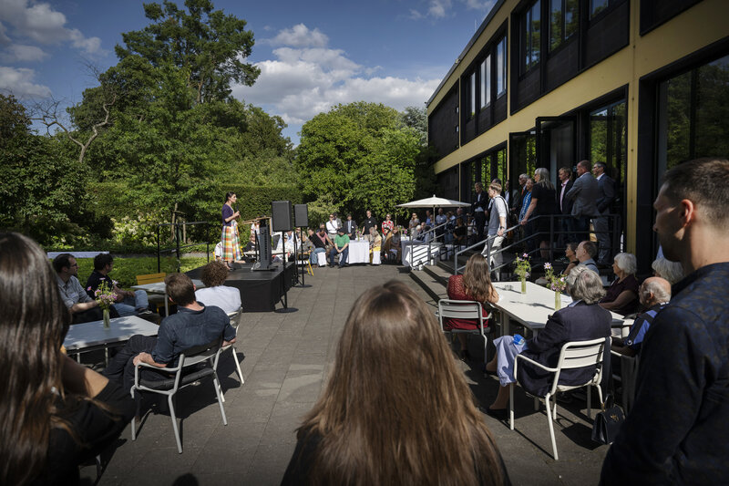 Im Garten der Akademie sitzen Männer und Frauen an Tischen. Eine Frau steht auf einer Bühne.