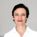 Eine Frau schaut lächelnd in die Kamera. Sie trägt eine weiße Bluse.