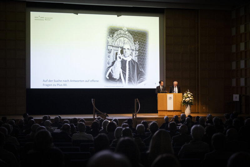 Zwei Männer stehen auf einer Bühne. Auf der Leinwand hinter ihnen ist ein Foto von Papst Pius XII. eingeblendet. 