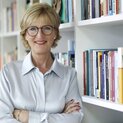 Frau mit verschränkten Armen steht lächelnd vor einem Bücherregal.