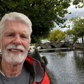 Ein Mann mit Bart steht unter einem Baum an einem Kanal. Im Hintergrund ist eine Brücke.