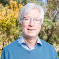 Ein Mann mit Brille steht lächelnd vor blühenden Strauchern.