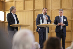 Drei Männer stehen auf einem Podium. Der mittlere spricht in ein Mikrofon. 