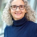 Eine Frau mit lockigem, langen Haar und Brille lächelt strahlend in die Kamera.