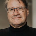 Ein Mann mit Brille lächelt in die Kamera. Er trägt einen schwarzen Rollkragenpullover.