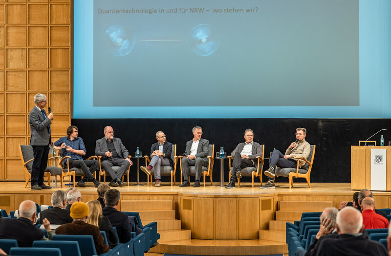 Auf einem Podium sitzen sechs Männer. An der Seite steht an siebter Mann mit einem Mikrofon.