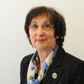 Eine Frau mit Brille lächelt in die Kamera. Sie trägt ein buntes Halstuch zu einer Anzugjacke.
