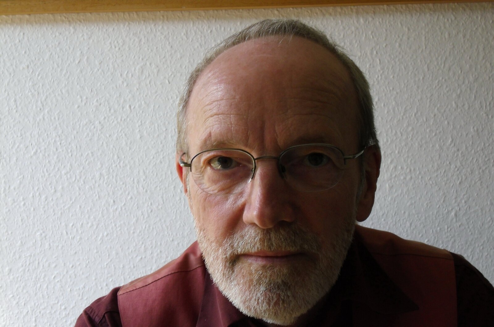 Ein Mann mit Brille und Bart sitzt vor einer weißen Tapete und schaut in die Kamera.