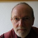 Ein Mann mit Brille und Bart sitzt vor einer weißen Tapete und schaut in die Kamera.
