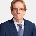 Ein Mann mit Brille schaut freundlich in die Kamera. Er trägt eine Krawatte und eine Anzugjacke.