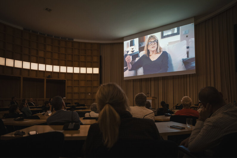 Ein Vortragsraum. Auf einer großen Leinwand ist eine Frau zu sehen.