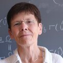 Eine Frau mit kurzem Haar und randloser Brille steht vor einer Tafel mit mathematischen Formeln. Sie schaut freundlich in die Kamera.