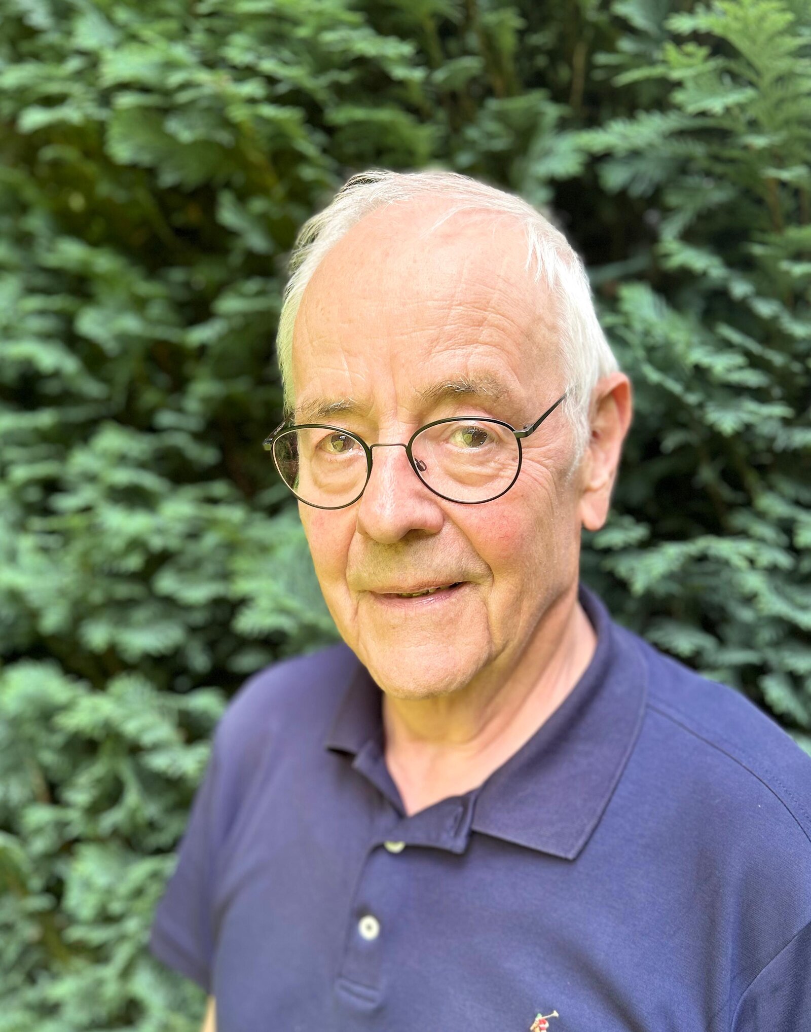 Ein Mann mit Brille steht lächelnd vor einem Strauch.