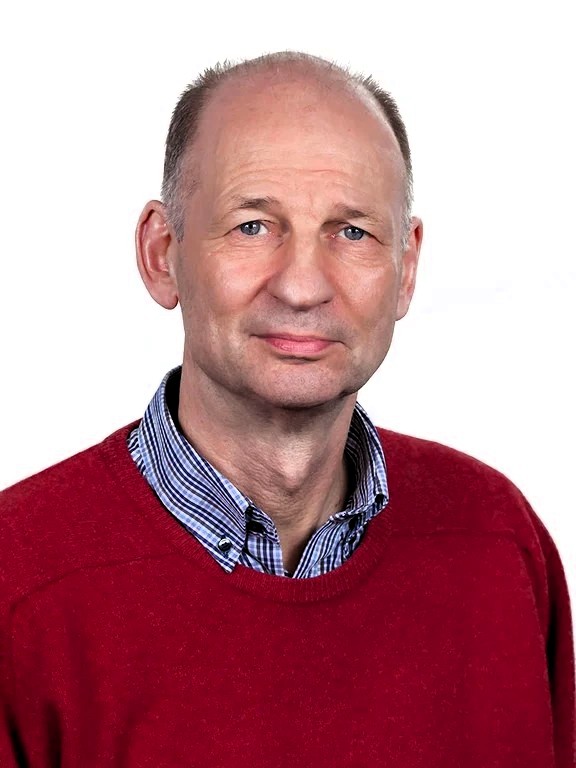 Ein Mann schaut freundlich in die Kamera. Er trägt ein kariertes Hemd unter einem roten Pullover.