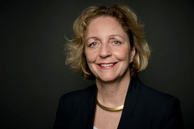 Eine Frau lächelt in die Kamera. Sie trägt einen Blazer und eine goldene Halskette.