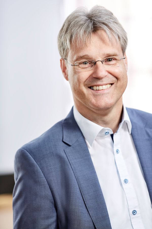 Ein Mann mit randloser, viereckiger Brille lächelt strahlend in die Kamera.