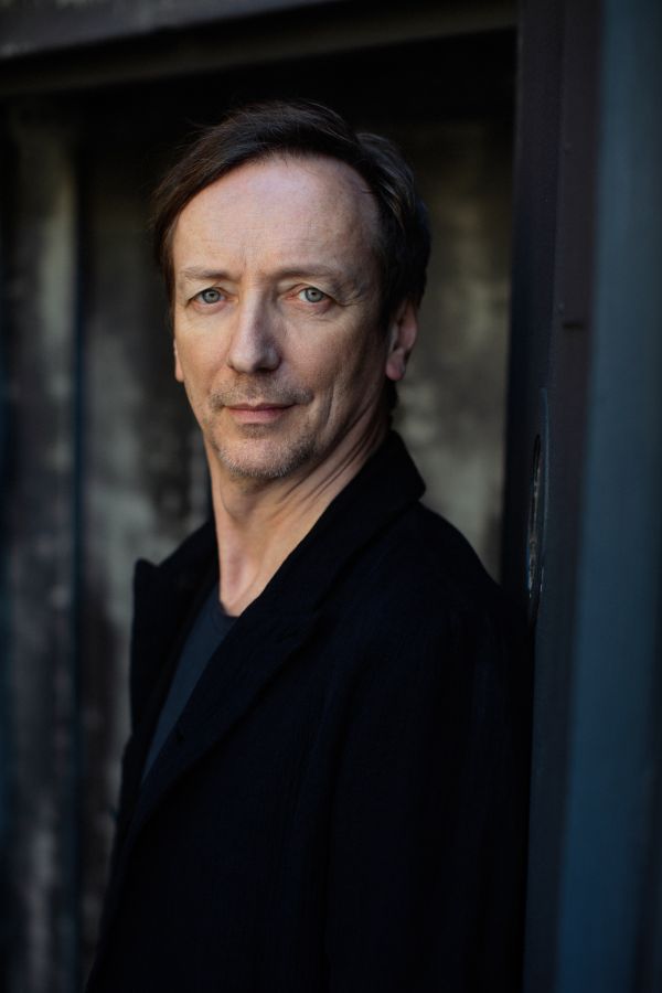 Ein Mann steht vor einer dunklen Wand. Er schaut freundlich in die Kamera.