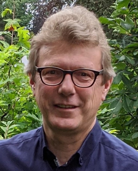 Ein Mann mit Brille steht vor einem Strauch und lächelt in die Kamera.