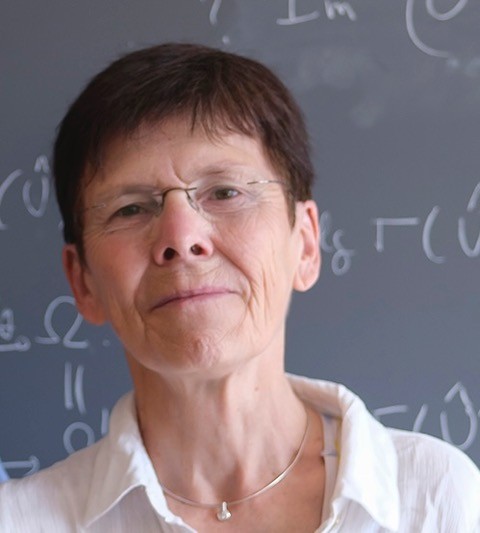 Eine Frau mit kurzem Haar und randloser Brille steht vor einer Tafel mit mathematischen Formeln. Sie schaut freundlich in die Kamera.