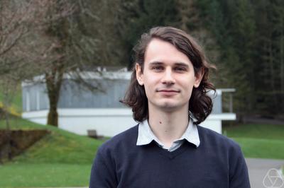 Ein Mann mit langen dunklen Haaren steht in einem Garten und sieht lächelnd in die Kamera. 
