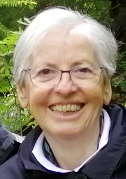 Eine Frau mit Brille steht vor Sträuchern und lächelt strahlend in die Kamera.