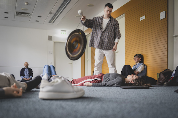 Männer und Frauen liegen auf dem Boden. In der Mitte des Raumes steht ein Mann, der einen großen Gong in der Hand hält.